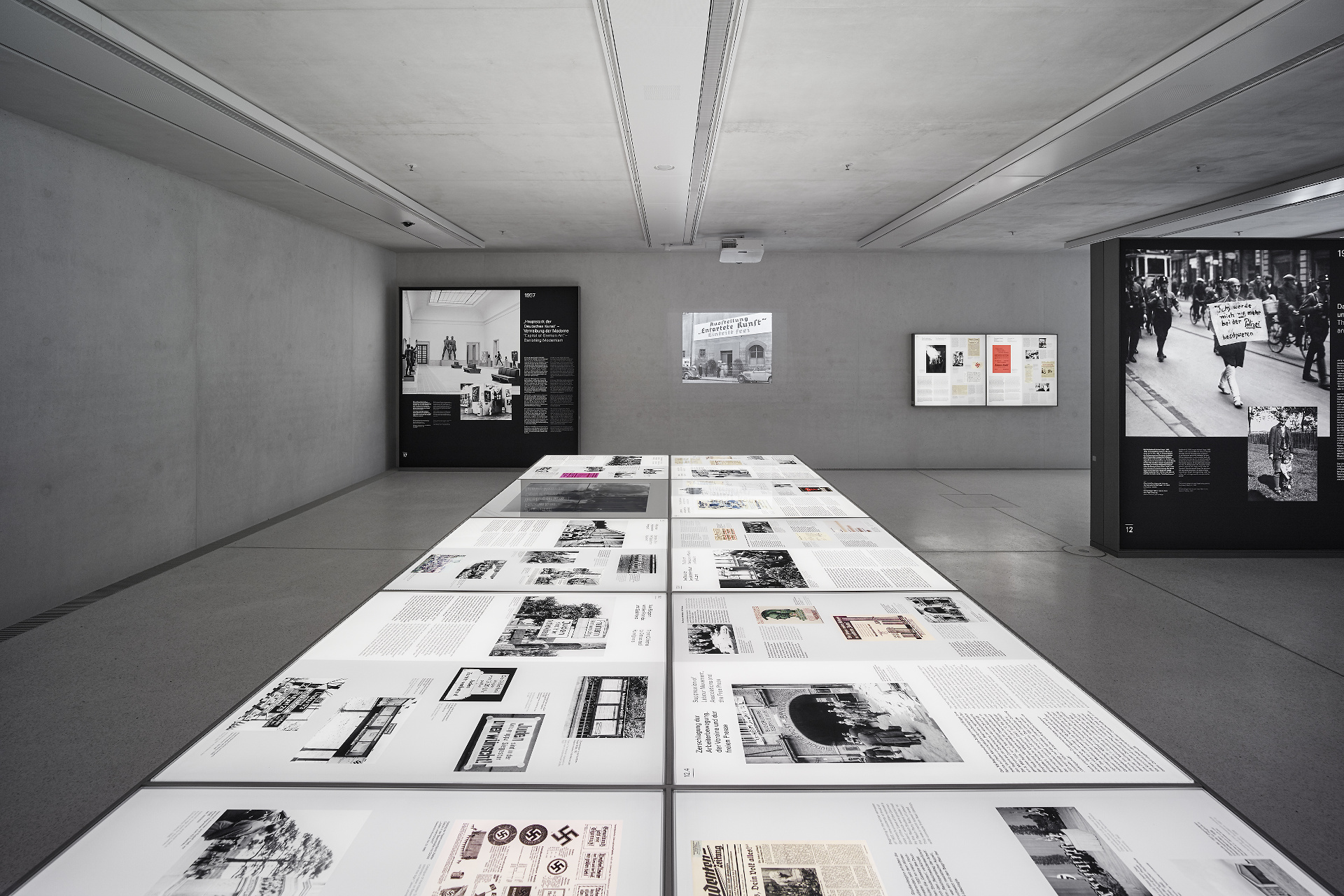 窓のない大きな部屋に並べられた複数の展示台と、文章や写真が印刷された大型の展示パネル。壁には映像が映されている。